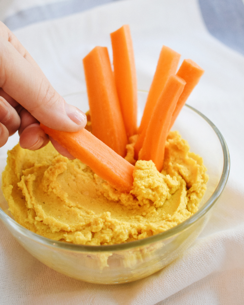 Carrot sticks shown being dipped in orange hummus